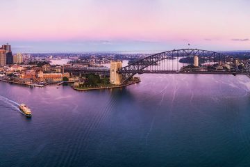 Pink sunrise over Sydney harbour, the harbour bridge and city CBDs on both shores in wide aerial panorama over calm waters disturbed by ferries and boats.